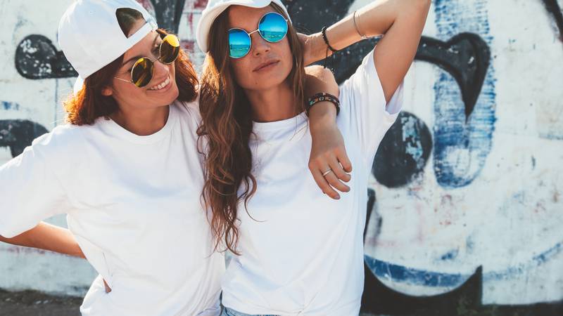 Two models wearing plain white t-shirts and hipster sunglasses posing against street wall. Teen urban clothing style, same look. Mockup for tshirt print store.